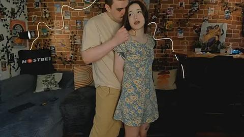 Media: Video of a young, slim, fair-skinned woman with long brown hair in a floral dress, being hugged from behind by a taller man with short dark hair in a beige shirt and khaki pants. Background features a brick wall with framed photos and a blue couch.