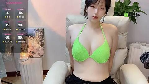 Media: A video of an Asian woman with fair skin and dark hair, wearing a neon green bikini, sitting in a white armchair in a cozy room with a potted plant.