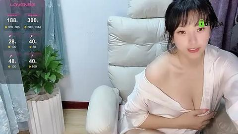 Media: Video of an East Asian woman with straight black hair, wearing a white satin robe, sitting on a cream-colored leather chair in a modern, minimalist living room.