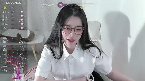 Media: Video of an East Asian woman with long black hair, glasses, and a white shirt, smiling, sitting in a modern room with white walls, a chair, and a monitor displaying game data.