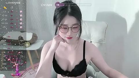 Media: Video of an East Asian woman with fair skin, wearing glasses and black lace lingerie, sitting on a couch. Background includes a gray wall, a cat statue, and a virtual reality headset.