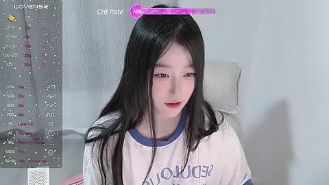 Media: Video of a young East Asian woman with long, straight black hair, fair skin, and red lipstick, wearing a white t-shirt with blue trim, sitting on a white chair in a minimalist room.