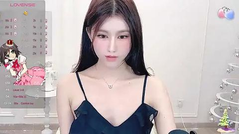 Media: Video of an Asian woman with long black hair, fair skin, and small breasts, wearing a black spaghetti-strap top, standing in a modern bathroom with white walls and a calendar poster featuring a cartoon girl in a pink dress.