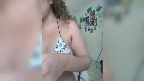 Media: A video of a topless woman with curly brown hair, wearing a floral bikini top, standing in a cluttered room with a white wall covered in colorful posters.