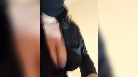 Media: Video of a woman with large breasts in a black top, showing cleavage, against a blurred background.