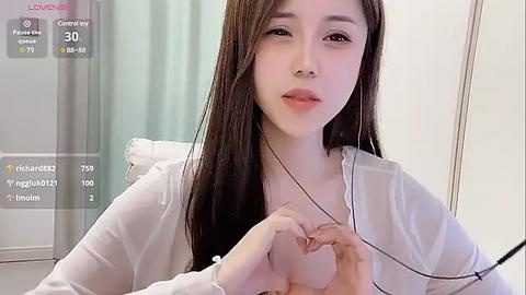 Media: A video of an Asian woman with long brown hair, fair skin, and light makeup, sitting in a room with white walls and a light green curtain. She is wearing a white cardigan and is focused on her phone, holding it with both hands.