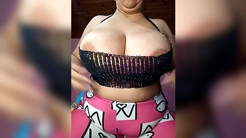 Media: Video of a woman with light skin, wearing a black, beaded bandeau top revealing large breasts, and pink pants with cartoon characters. Background is blurred.