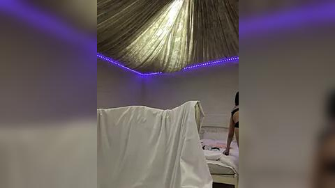 Media: Video of a modern, minimalist room with a white bed draped in a white sheet, a person in a black shirt standing by, and a ceiling with a circular, textured, silver fabric design illuminated by blue LED lights.