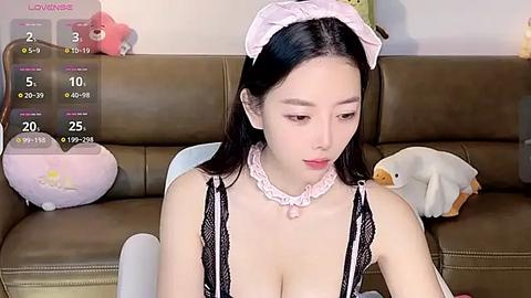 Media: Video of an East Asian woman in a black lace maid outfit, sitting on a brown couch, with a pink headband, fluffy white pillow, and plush toys in the background.