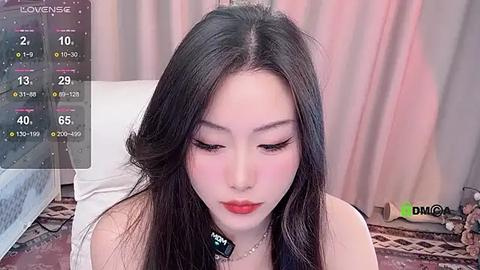 Media: Video of a young East Asian woman with long black hair, fair skin, and red lipstick, sitting indoors, wearing a black top and necklace, against a soft pink curtain background.