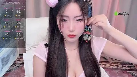 Media: A video of an East Asian woman with long black hair, wearing a pink headband, a floral hair accessory, and a light pink top, sitting indoors with a white couch and patterned carpet.