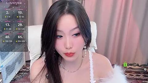 Media: Video of a young East Asian woman with long, straight black hair, wearing a white off-shoulder lace dress with feather details, in a bedroom setting with pink curtains and a white pillow.