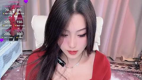 Media: A video of an East Asian woman with long black hair, fair skin, and red lipstick, wearing a red top, sitting on a bed. Background includes a white pillow and pink curtains.