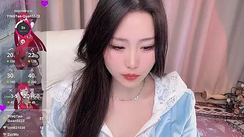 Media: Video of a young East Asian woman with long black hair, wearing a light blue hoodie, sitting on a white leather chair. The background shows a soft pink curtain and a decorative rug. Live streaming overlay with chat messages.