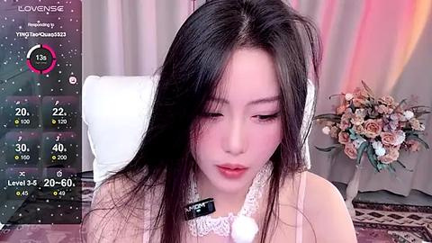 Media: A video of an Asian woman with long black hair, wearing a white lace top, sitting in front of a floral arrangement and a white chair, with a virtual background of a cozy room.