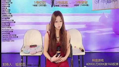 Media: Video of an Asian woman with long brown hair in a red dress, holding a microphone, seated between two white chairs with stuffed toys on them. Background features Chinese text and a purple gradient.