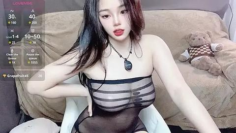 Media: A video of an Asian woman with long black hair, wearing a sheer black top, sitting on a light-colored couch. A teddy bear is beside her, and the background includes a virtual live streaming screen with metrics.