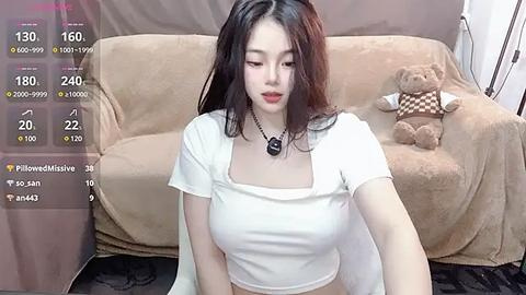 Media: A video of an East Asian woman with long black hair and fair skin, wearing a white crop top, sitting on a beige couch with a teddy bear, in a cozy living room.