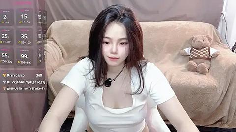 Media: A video of an East Asian woman with long black hair, wearing a white shirt and necklace, sitting on a beige couch with a teddy bear, in a cozy, indoor setting.