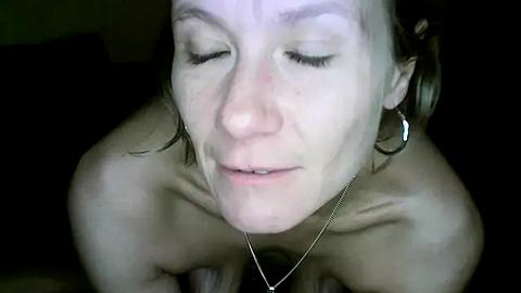 Media: Video of a Caucasian woman with light skin, closed eyes, and a neutral expression, wearing a silver necklace and large hoop earrings, taken in dim lighting.