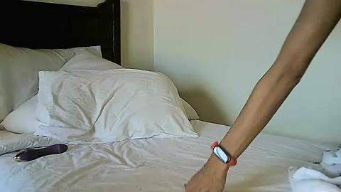 Media: Video of a person in a white shirt and black wristwatch standing next to an unmade bed with white sheets and pillows, with a purple vibrator on the bed.