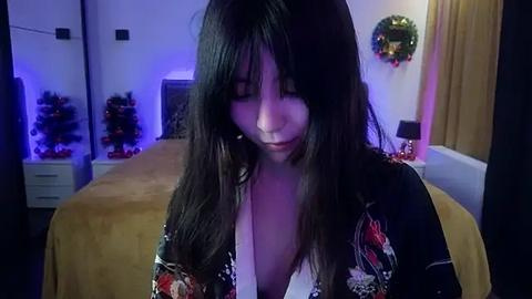 Media: A video of a young woman with long black hair and pale skin, wearing a low-cut black kimono, standing in a dimly lit bedroom adorned with Christmas decorations.