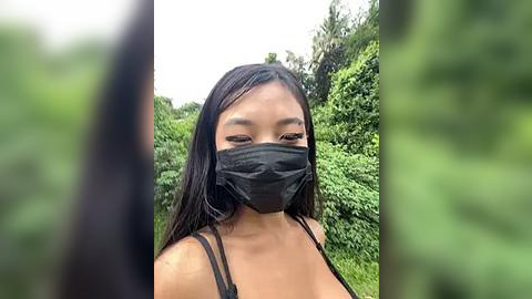 Media: Video of a young Asian woman with long black hair, wearing a black face mask and a black spaghetti-strap top, standing in a lush, green garden. The image is slightly blurred, emphasizing her face and greenery in the background.