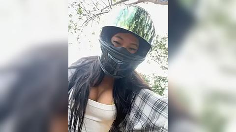 Media: A video of a woman with medium brown skin and long black hair, wearing a reflective green helmet, black mask, white tank top, and a plaid jacket. The background features blurred greenery.