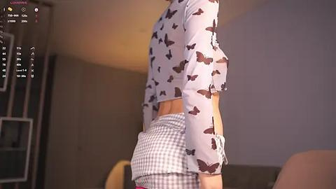 Media: Video of a slender woman with fair skin, wearing a light purple crop top with butterfly print and checkered shorts, standing indoors near a bookshelf.