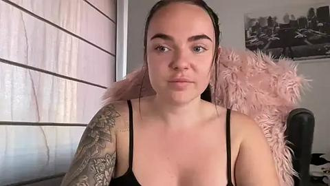 Media: A video of a fair-skinned woman with brown hair, tattoos on her right arm, wearing a black spaghetti-strap top, seated in a room with a pink furry chair, light-colored walls, and a cityscape print on the wall.