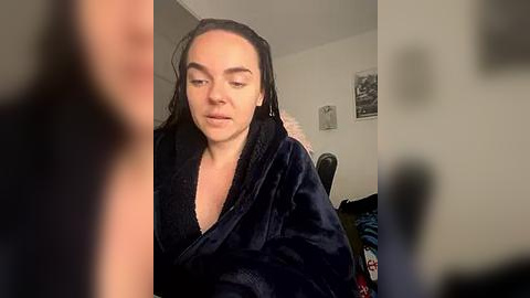 Media: Video of a light-skinned woman with dark hair in a robe, appearing to be in a bedroom with blurred background items, including a framed picture and clothes.