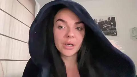 Media: Video of a woman in a dark robe, wearing black makeup and a confused expression. The background features a white wall with a framed black-and-white video and a light-colored wall panel.