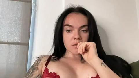 Media: Video of a young woman with fair skin, long black hair, and green eyes, wearing a red lace bra, resting her chin on her hand, with a tattoo on her left forearm. Background shows a frosted glass window and white wall.