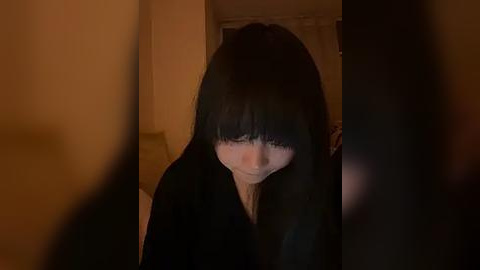 Media: A video of a young woman with long black hair, wearing a black top, standing indoors in a dimly lit room. The background is blurred, and her face is partially obscured by her hair.