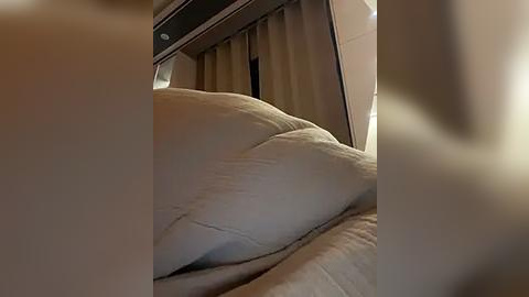 Media: Video of a plush, light beige sofa with soft cushions, positioned in a modern, dimly lit room with beige curtains and a dark, recessed ceiling light.