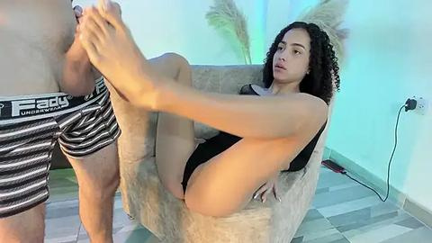 Media: A video of a woman with curly hair, wearing a black bodysuit, being massaged by a man in black and white striped underwear on a light gray chair.