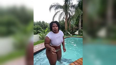 Media: Video of a plus-sized woman with dark skin, wearing a white crop top and green bikini bottoms, standing in a swimming pool. Background includes lush greenery and palm trees.