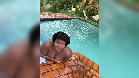 Media: A video shows a smiling Black woman with short curly hair and medium breasts, wearing a white bikini top, sitting on the edge of a turquoise pool with a brick-tiled surround. The background features lush greenery and a hammock.