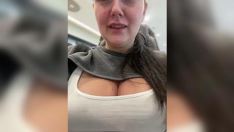 Media: Video of a woman with fair skin and brown hair, wearing a gray sweater revealing her cleavage. Background is blurred, showing a modern room with white walls and ceiling lights.