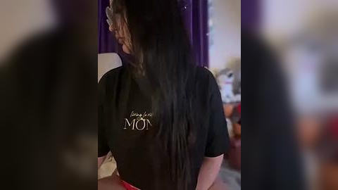 Media: Video of a young woman with long, straight black hair, wearing glasses, a black T-shirt with white text, and a red apron. Background shows purple curtains and a blurred room.