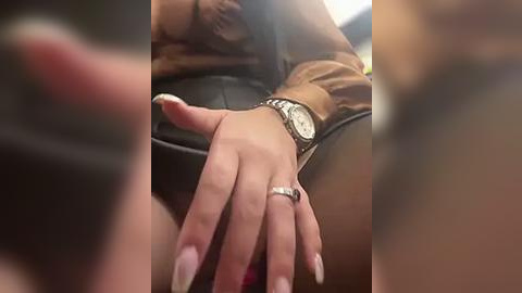 Media: Video of a woman's hand with long, manicured nails, wearing a silver watch and ring, resting on a black leather surface. Blurred background suggests an indoor setting.