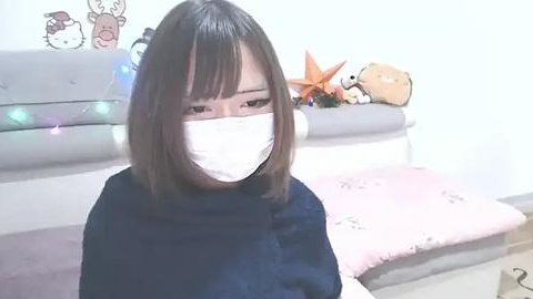 Media: Video of a young Asian woman with shoulder-length black hair and a surgical mask, wearing a black turtleneck sweater. She sits on a pink cushioned bench in a room with white walls and decorative pillows featuring cartoon characters.