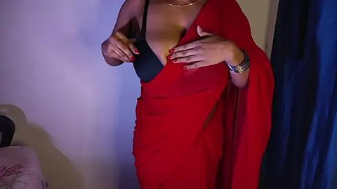 Media: Video of a woman with medium brown skin, wearing a red saree with a black bra visible. Her nails are painted red, and she has a gold bracelet on her wrist. The background features a dark blue curtain and a light-colored wall.