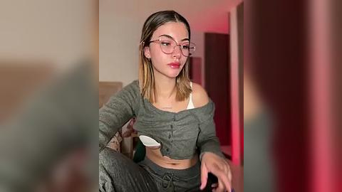 Media: Video of a young woman with light skin, straight brown hair, and round glasses, wearing a gray off-shoulder crop top and loose pants, seated on a couch in a dimly lit room with red lighting.