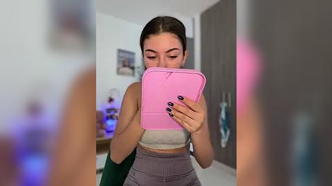 Media: A video of a young woman with fair skin, dark hair, and medium build, wearing a pink sweatshirt and high-waisted gray leggings, taking a selfie with a pink case, in a modern, minimally decorated room.