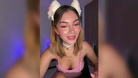 Media: Video of a young Asian woman with light skin, wearing a pink top, cat ears, and a choker, smiling at the camera in a dimly lit room.