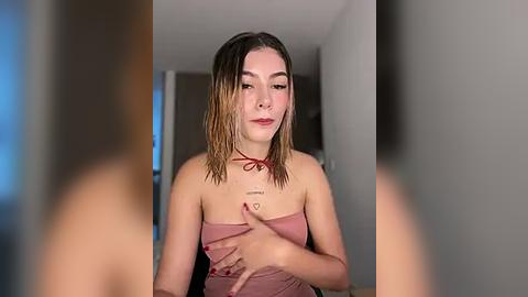 Media: Video of a young woman with wet, shoulder-length brown hair, wearing a strapless pink top, standing in a modern, minimalist room with neutral walls and a blurred, indistinct background.