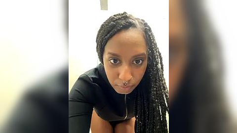 Media: Video of a Black woman with medium-brown skin, wearing black hair in braids, a black top, and a septum ring, leaning forward, looking directly at the camera, with blurred background.