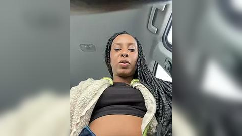 Media: Video of a young Black woman with braided hair, wearing a black crop top and a white knitted jacket, seated in a car with gray interior.