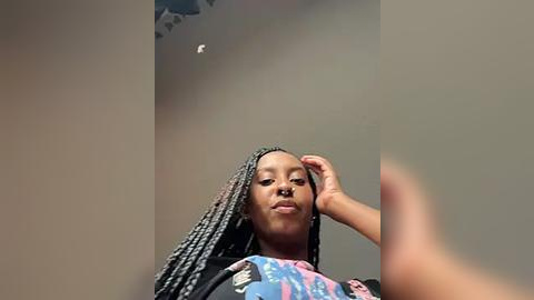 Media: Video of a young Black woman with medium-brown skin, wearing a black and colorful graphic t-shirt, with braided hair, posing in a room with neutral-colored walls and a ceiling light fixture.